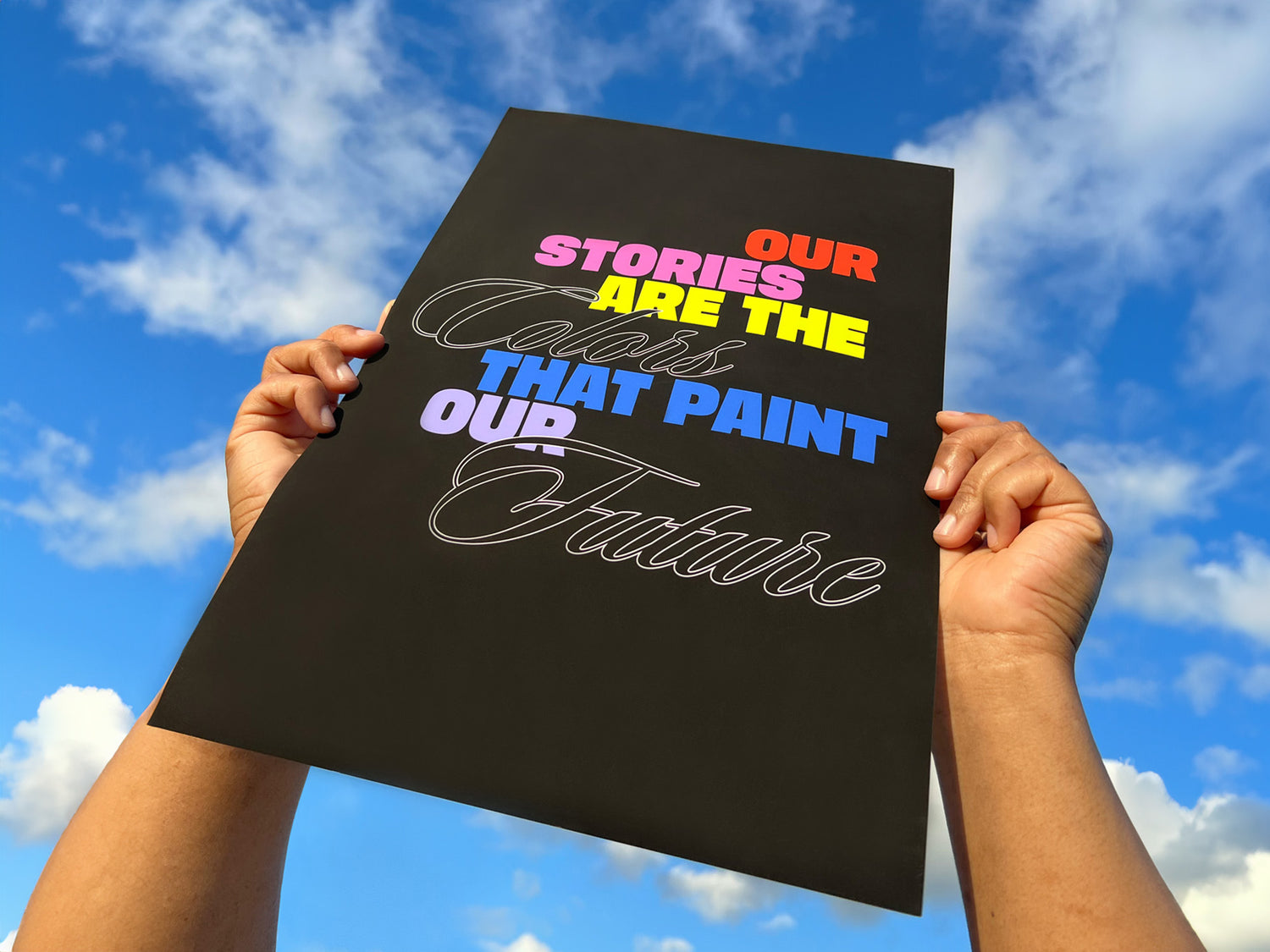 Hands hold a Partner poster in the air, the poster reads "Our Stories Are The Colors That Paint Our Future"